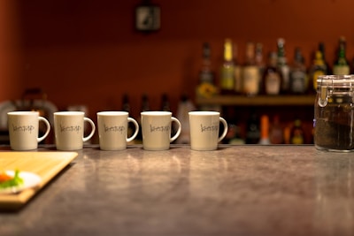 A row of five white mugs with a logo are lined up on a countertop in a bar setting. Behind them, a shelf filled with various bottles of alcohol is visible. To the right, a clear jar containing a dark substance is present. The background features a warm, ambient lighting that contrasts with the muted tones of the countertop.