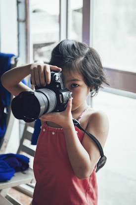 A young child in a pink dress holding a large camera, looking intently through the viewfinder. The setting appears to be indoors with a softly lit background and a partially open window, contributing to a calm atmosphere.