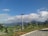 A scenic view featuring a well-maintained park with neatly arranged plants and bushes in the foreground. Solar-powered street lights are evenly spaced along a paved path. In the background, lush green hills are partially shrouded by low-lying clouds under a vibrant blue sky.