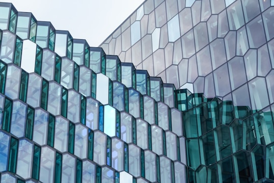 Glass window facade consisting of numerous hexagonal glass panels arranged in a geometric pattern, reflecting the sky and surroundings. Some glass panels are tinted blue and green, adding a variation in color.