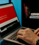 A person wearing a smartwatch types on a laptop with a website about web development displayed on the screen. The background includes a bookshelf with some books and a decorative horse sculpture.