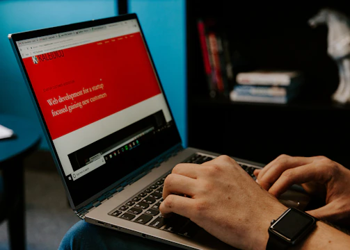 A person wearing a smartwatch types on a laptop with a website about web development displayed on the screen. The background includes a bookshelf with some books and a decorative horse sculpture.