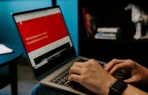 A person wearing a smartwatch types on a laptop with a website about web development displayed on the screen. The background includes a bookshelf with some books and a decorative horse sculpture.
