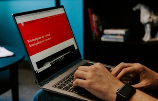 A person wearing a smartwatch types on a laptop with a website about web development displayed on the screen. The background includes a bookshelf with some books and a decorative horse sculpture.