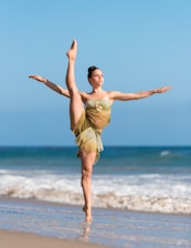A dancer is performing an elegant high kick on a sandy beach. The ocean waves are gently rolling in the background under a clear blue sky. The dancer wears a flowing, earth-toned dress and exudes grace and strength.