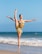 A dancer is performing an elegant high kick on a sandy beach. The ocean waves are gently rolling in the background under a clear blue sky. The dancer wears a flowing, earth-toned dress and exudes grace and strength.