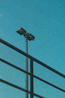 A tall pole with two floodlights mounted on top is visible against a clear blue sky. A metal mesh fence runs diagonally across the foreground.