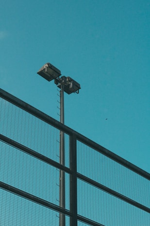 A tall pole with two floodlights mounted on top is visible against a clear blue sky. A metal mesh fence runs diagonally across the foreground.