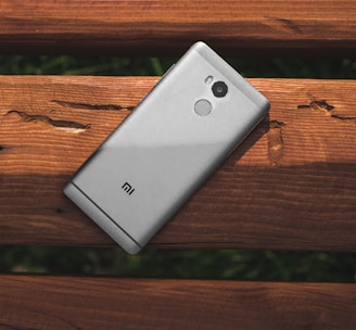 A silver smartphone with a camera and fingerprint sensor is resting on a weathered wooden surface with visible texture and grain.