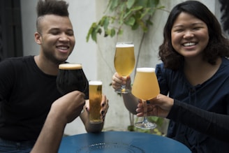 A group of friends enjoying a gathering, holding up different types of beer glasses in a toast. Two people are prominently smiling, and the setting appears to be casual and outdoors with some greenery in the background.