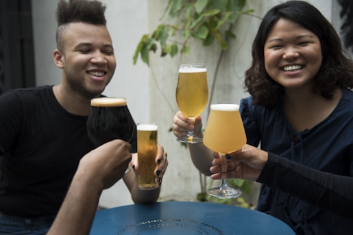 A group of friends enjoying a gathering, holding up different types of beer glasses in a toast. Two people are prominently smiling, and the setting appears to be casual and outdoors with some greenery in the background.