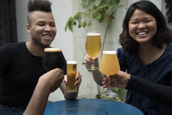 A group of friends enjoying a gathering, holding up different types of beer glasses in a toast. Two people are prominently smiling, and the setting appears to be casual and outdoors with some greenery in the background.