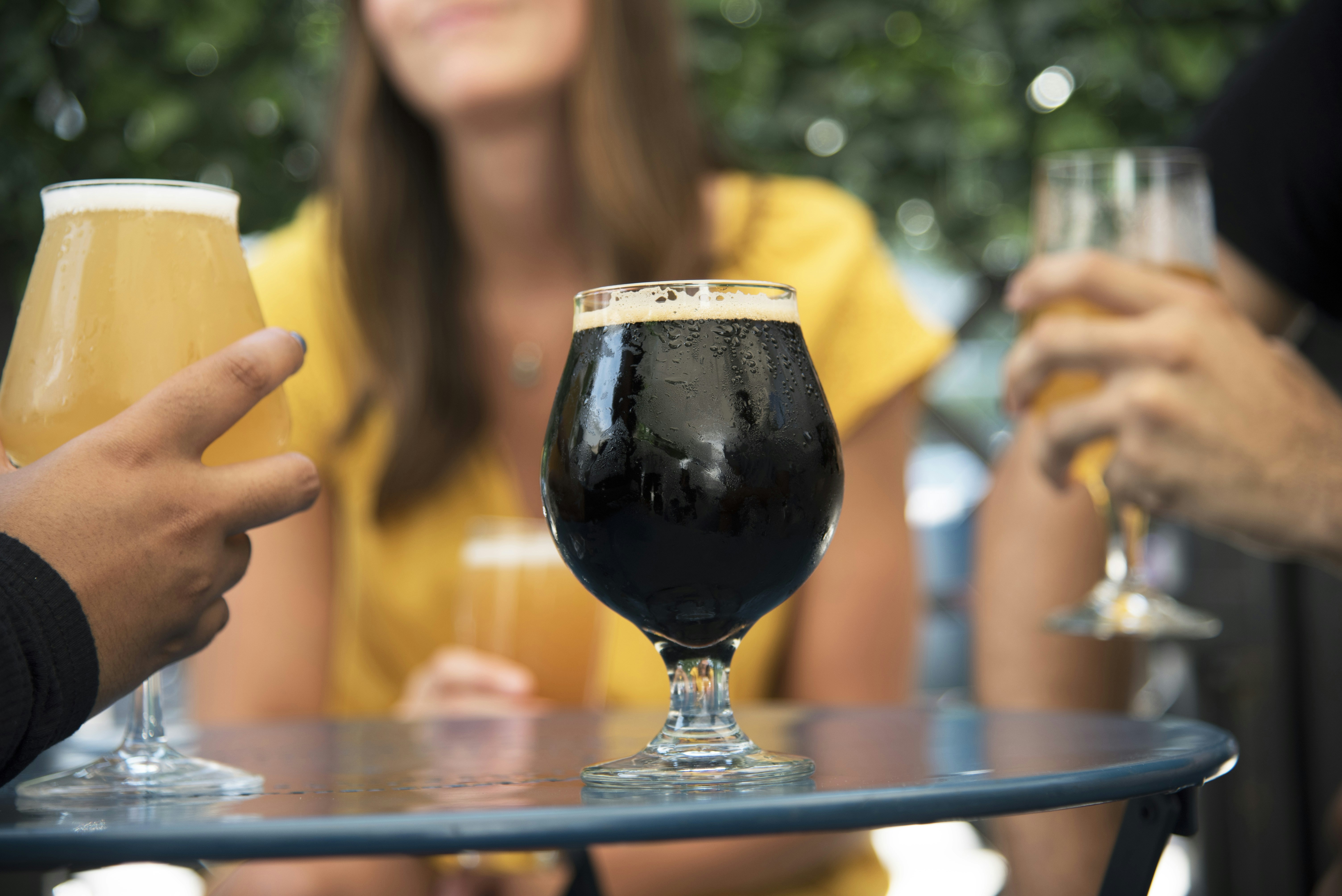 A beer glass on a table filled with a stout with people drinking lighter beer in the background.