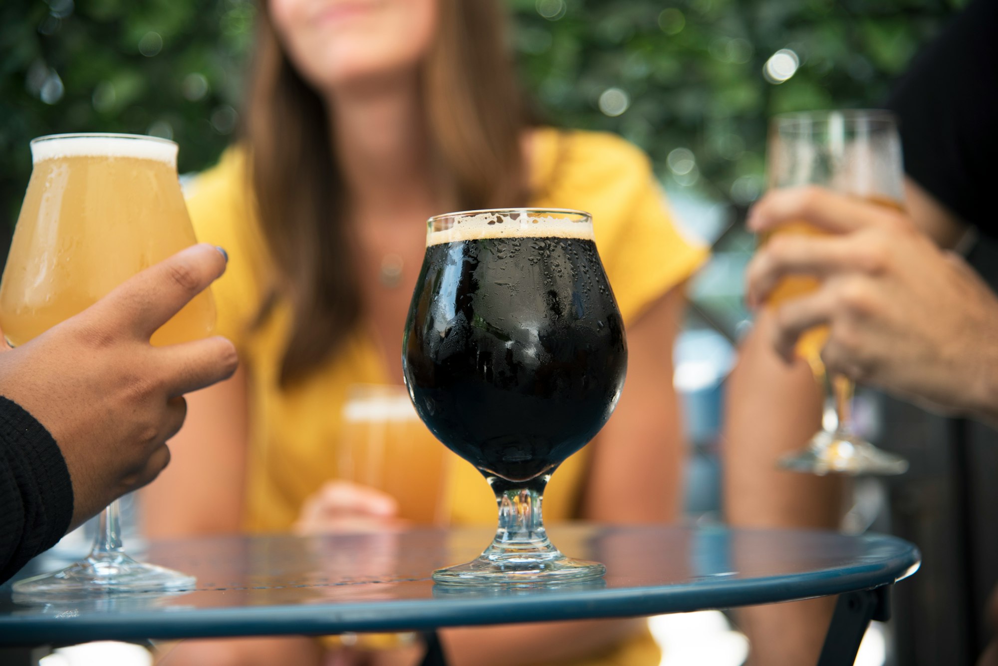 A beer glass on a table filled with a stout with people drinking lighter beer in the background.