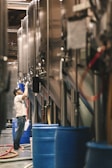 A person wearing a cap and a white t-shirt is working in a brewery or industrial setting, surrounded by large stainless steel tanks. Blue barrels and various hoses are visible, suggesting an active production environment.