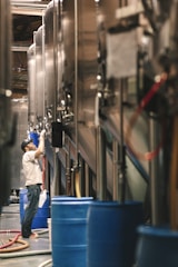 A person wearing a cap and a white t-shirt is working in a brewery or industrial setting, surrounded by large stainless steel tanks. Blue barrels and various hoses are visible, suggesting an active production environment.