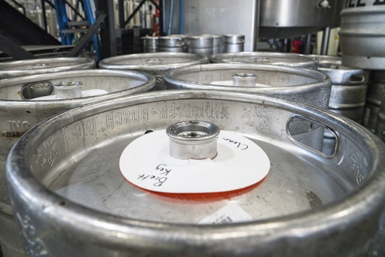 Several steel kegs stacked together in a brewery setting. The kegs have branding engraved on them, and one keg has a white label with handwritten text.