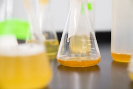 A close-up of several laboratory flasks containing a yellow liquid. The flasks vary in size and are positioned on a dark surface, creating an environment suggestive of a scientific or experimental setting.
