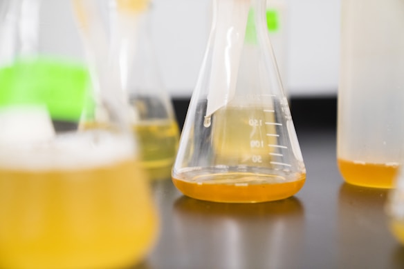 A close-up of several laboratory flasks containing a yellow liquid. The flasks vary in size and are positioned on a dark surface, creating an environment suggestive of a scientific or experimental setting.
