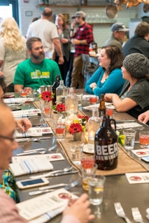 A table is set for a gathering with a variety of drinks and flowers as a centerpiece. People are seated and standing around, engaging in conversation in a relaxed atmosphere. The setting appears to be a restaurant or bar, with a rustic and cozy decor.