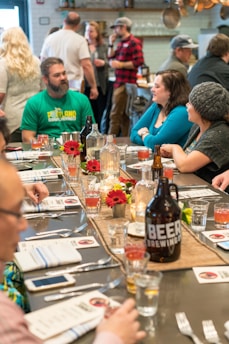 A table is set for a gathering with a variety of drinks and flowers as a centerpiece. People are seated and standing around, engaging in conversation in a relaxed atmosphere. The setting appears to be a restaurant or bar, with a rustic and cozy decor.