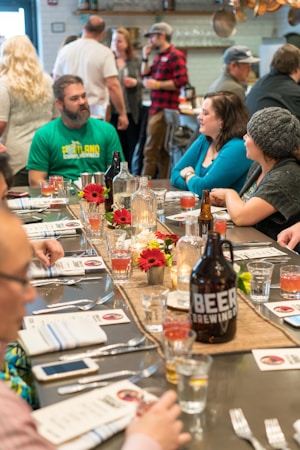 A table is set for a gathering with a variety of drinks and flowers as a centerpiece. People are seated and standing around, engaging in conversation in a relaxed atmosphere. The setting appears to be a restaurant or bar, with a rustic and cozy decor.