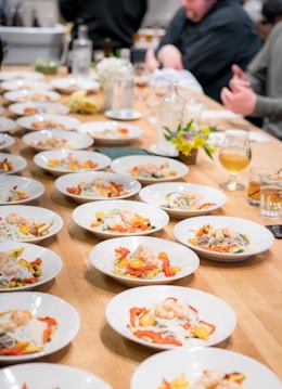 Multiple white plates arranged neatly on a wooden table, each containing a colorful dish of shrimp, sliced red and yellow bell peppers, and noodles. The table is in a communal dining setting with several people seated in the background, engaging in conversation. Some drinks, flowers, and glass bottles are also part of the table setting.