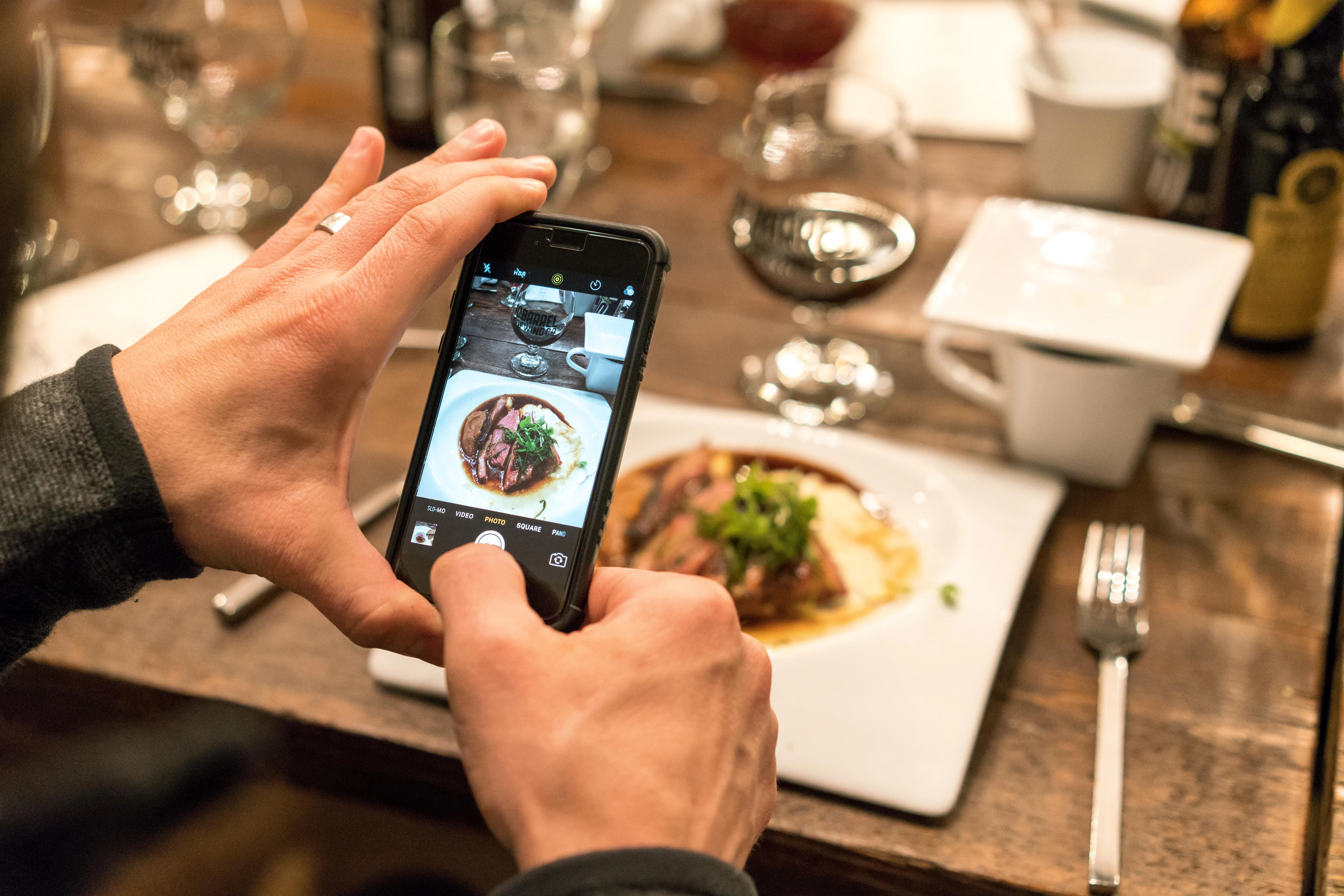 Hands holding a black iPhone over a restaurant table, photographing a plate of meat with green herbs, wine glasses in the background