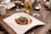 A gourmet dish is elegantly presented on a uniquely shaped white plate, featuring seared meat topped with fresh greens, accompanied by a rich sauce and a side of creamy mashed potatoes. The setup is on a rustic wooden table, surrounded by various glasses filled with different beverages, creating a cozy dining atmosphere.