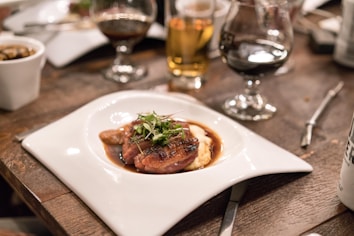 A gourmet dish is elegantly presented on a uniquely shaped white plate, featuring seared meat topped with fresh greens, accompanied by a rich sauce and a side of creamy mashed potatoes. The setup is on a rustic wooden table, surrounded by various glasses filled with different beverages, creating a cozy dining atmosphere.