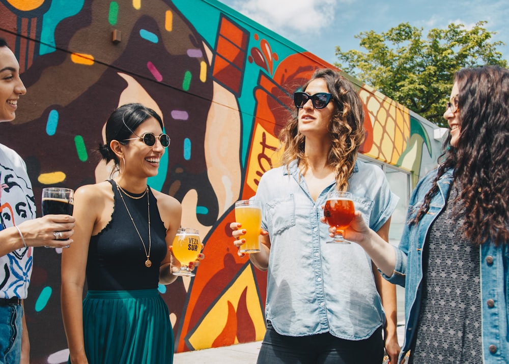 Cuatro mujeres hablando entre sí sosteniendo vasos transparentes mientras están de pie cerca de una pared con grafitis durante el día