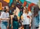 A group of four friends is gathered outdoors, enjoying colorful drinks. They are standing in front of a vibrant mural featuring abstract patterns. The friends are laughing and appear to be in a cheerful mood. They are wearing casual summer clothing, including sunglasses, and are holding different colored drinks.