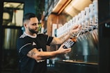 A man with tattoos is filling a glass bottle with a beverage from a row of taps at a bar or brewery. He wears a black shirt with white details. The background shows a stainless steel surface and some shelves with bottles, creating a casual and industrial atmosphere.