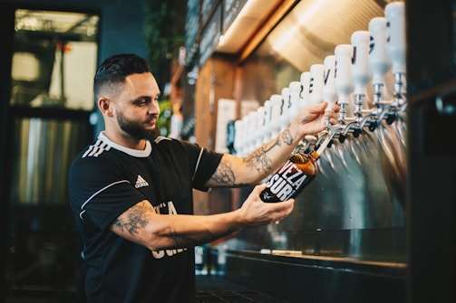 A man with tattoos is filling a glass bottle with a beverage from a row of taps at a bar or brewery. He wears a black shirt with white details. The background shows a stainless steel surface and some shelves with bottles, creating a casual and industrial atmosphere.