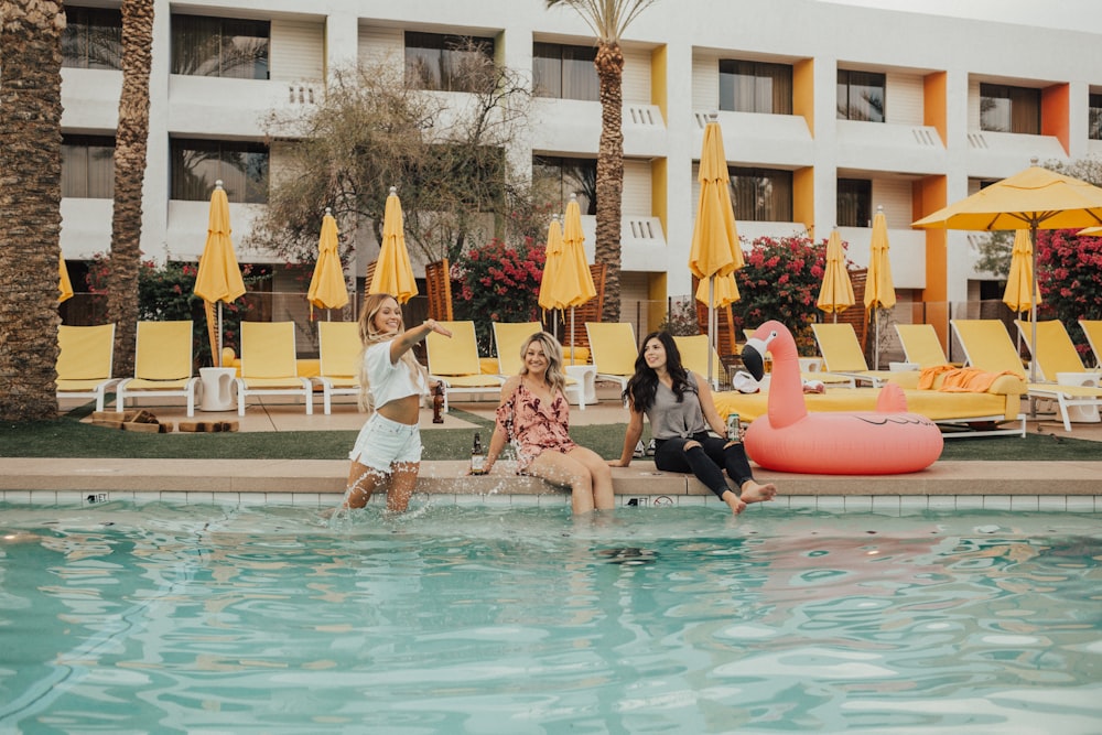 three women sitting beside flamingo floater near swimming pool during daytime