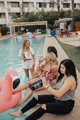 Four women are relaxing by a pool, with one on a large pink flamingo float in the water. They appear to be enjoying themselves, holding drinks, and chatting. The setting looks like a resort or a vacation spot with a bright, casual atmosphere.