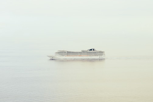 A large cruise ship is sailing through calm waters with a clear and minimalistic backdrop. The ship is mid-sized, featuring multiple decks and a sleek, modern design.