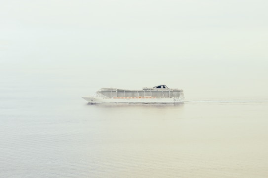 A large cruise ship is sailing through calm waters with a clear and minimalistic backdrop. The ship is mid-sized, featuring multiple decks and a sleek, modern design.