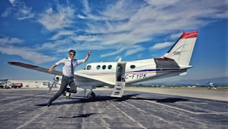 A person in pilot attire is captured in mid-air jump next to a small airplane stationed on an airport tarmac. The sky is clear with some clouds, and mountains are visible in the background. Other aircraft and airport facilities can be seen in the distance.