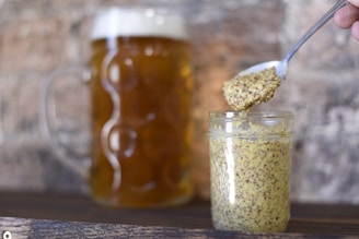 A glass jar filled with whole grain mustard sits on a wooden surface. A spoon, held by a hand, is lifted above the jar, carrying a small amount of the mustard. In the background, a large mug of beer with a frothy head is visible.