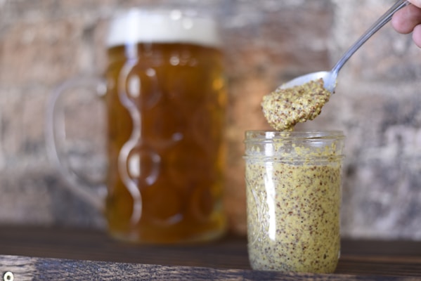 A glass jar filled with whole grain mustard sits on a wooden surface. A spoon, held by a hand, is lifted above the jar, carrying a small amount of the mustard. In the background, a large mug of beer with a frothy head is visible.