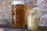 A glass jar filled with whole grain mustard sits on a wooden surface. A spoon, held by a hand, is lifted above the jar, carrying a small amount of the mustard. In the background, a large mug of beer with a frothy head is visible.