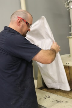 A person wearing safety glasses and a navy blue shirt is holding a large white sack, pouring its contents onto a piece of machinery. The focus is on the action of emptying the sack, with some industrial equipment in the background.