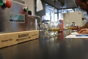 A laboratory setting features a line of glasses partially filled with a golden liquid, placed on a shiny black countertop. In the foreground, there is a cardboard box with 'KARBACH BREWING CO' written on it. To the side, a person is writing on a piece of paper with a pen, surrounded by various equipment and machinery.
