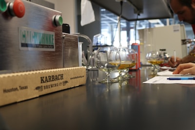 A laboratory setting features a line of glasses partially filled with a golden liquid, placed on a shiny black countertop. In the foreground, there is a cardboard box with 'KARBACH BREWING CO' written on it. To the side, a person is writing on a piece of paper with a pen, surrounded by various equipment and machinery.