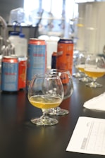 On a table, several snifter glasses partially filled with a golden liquid are placed alongside colorful beverage cans. There are some laboratory bottles in the background, and a paper with a chart or table is visible in the foreground.