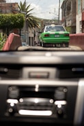 A green and white taxi drives down a rustic street with buildings and palm trees lining the sides. The image is viewed through a car windshield with a blurred dashboard in the foreground. It appears to be a bright day with a clear sky.