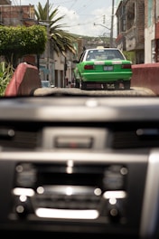 A green and white taxi drives down a rustic street with buildings and palm trees lining the sides. The image is viewed through a car windshield with a blurred dashboard in the foreground. It appears to be a bright day with a clear sky.