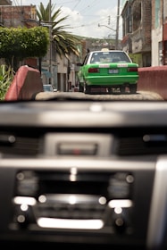 A green and white taxi drives down a rustic street with buildings and palm trees lining the sides. The image is viewed through a car windshield with a blurred dashboard in the foreground. It appears to be a bright day with a clear sky.