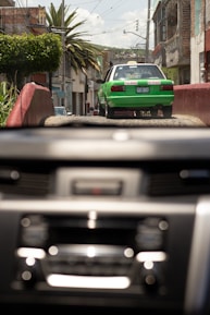 A green and white taxi drives down a rustic street with buildings and palm trees lining the sides. The image is viewed through a car windshield with a blurred dashboard in the foreground. It appears to be a bright day with a clear sky.